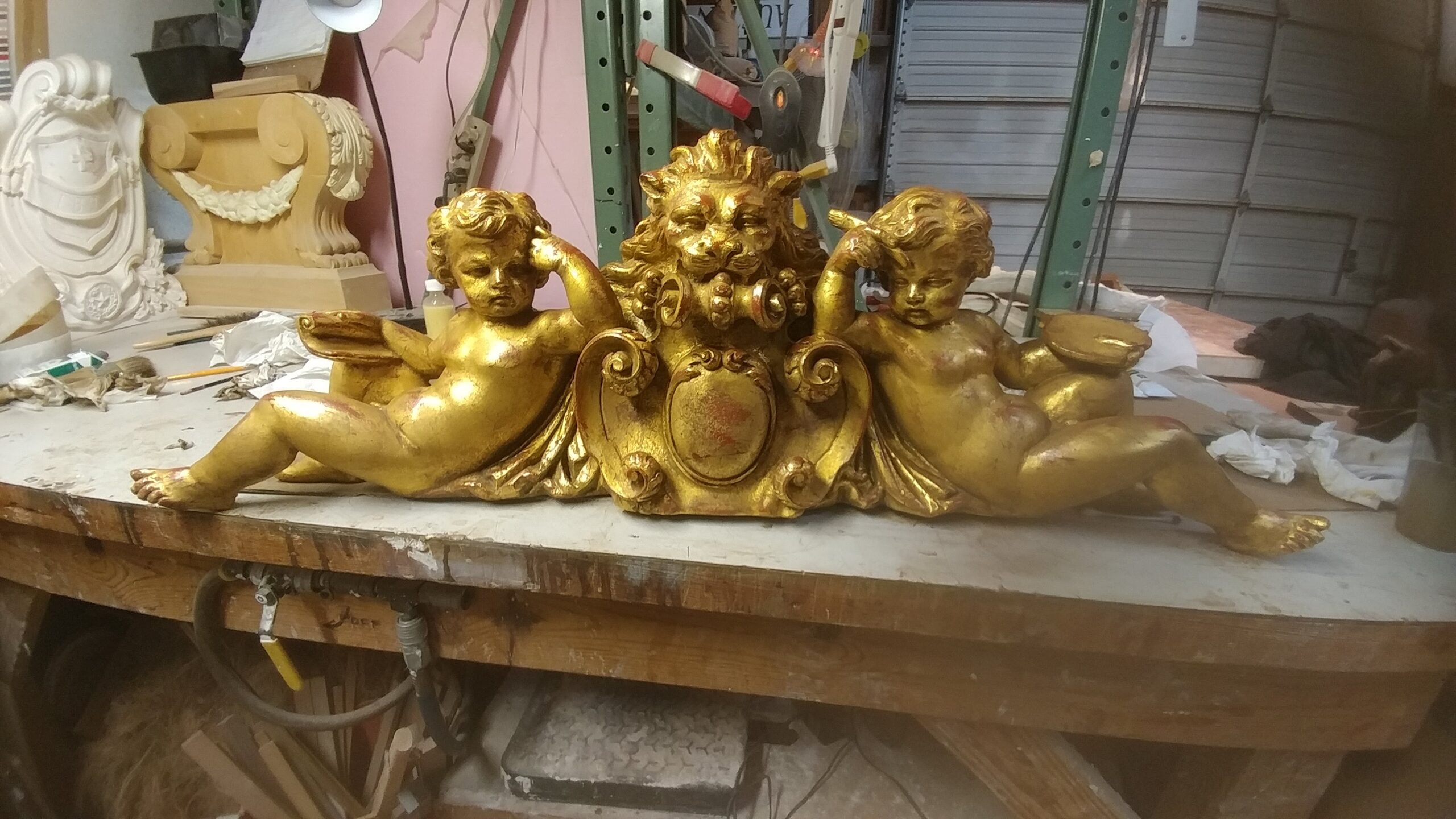 Gilded carved overmantel with cherubs and a lion mask resting on a workbench in a Dallas gilding workshop